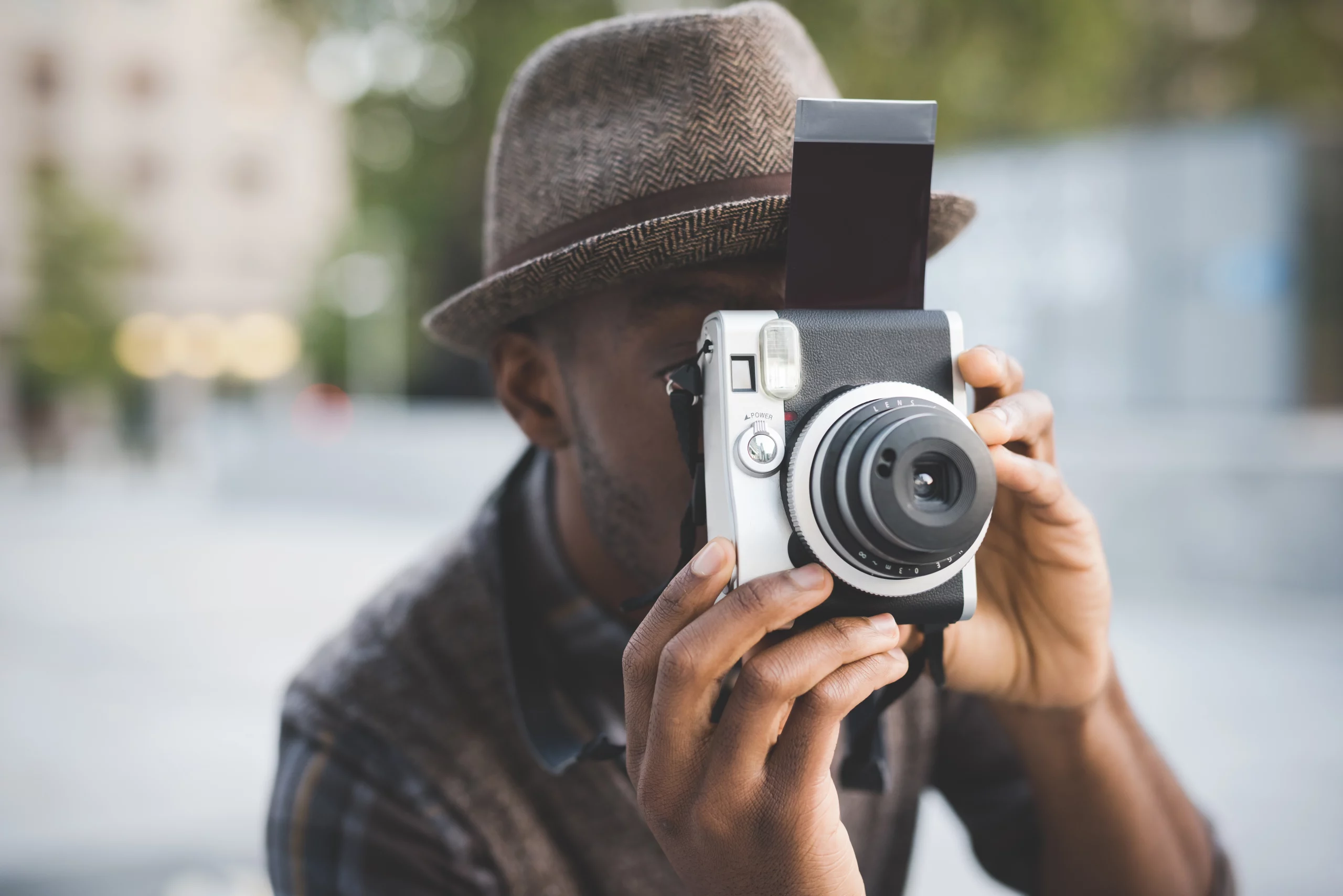 A man on vacation is taking a picture with an instax camera while wearing a hat.