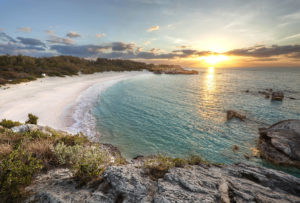 The sun is setting over a sandy beach and rocky cliffs.